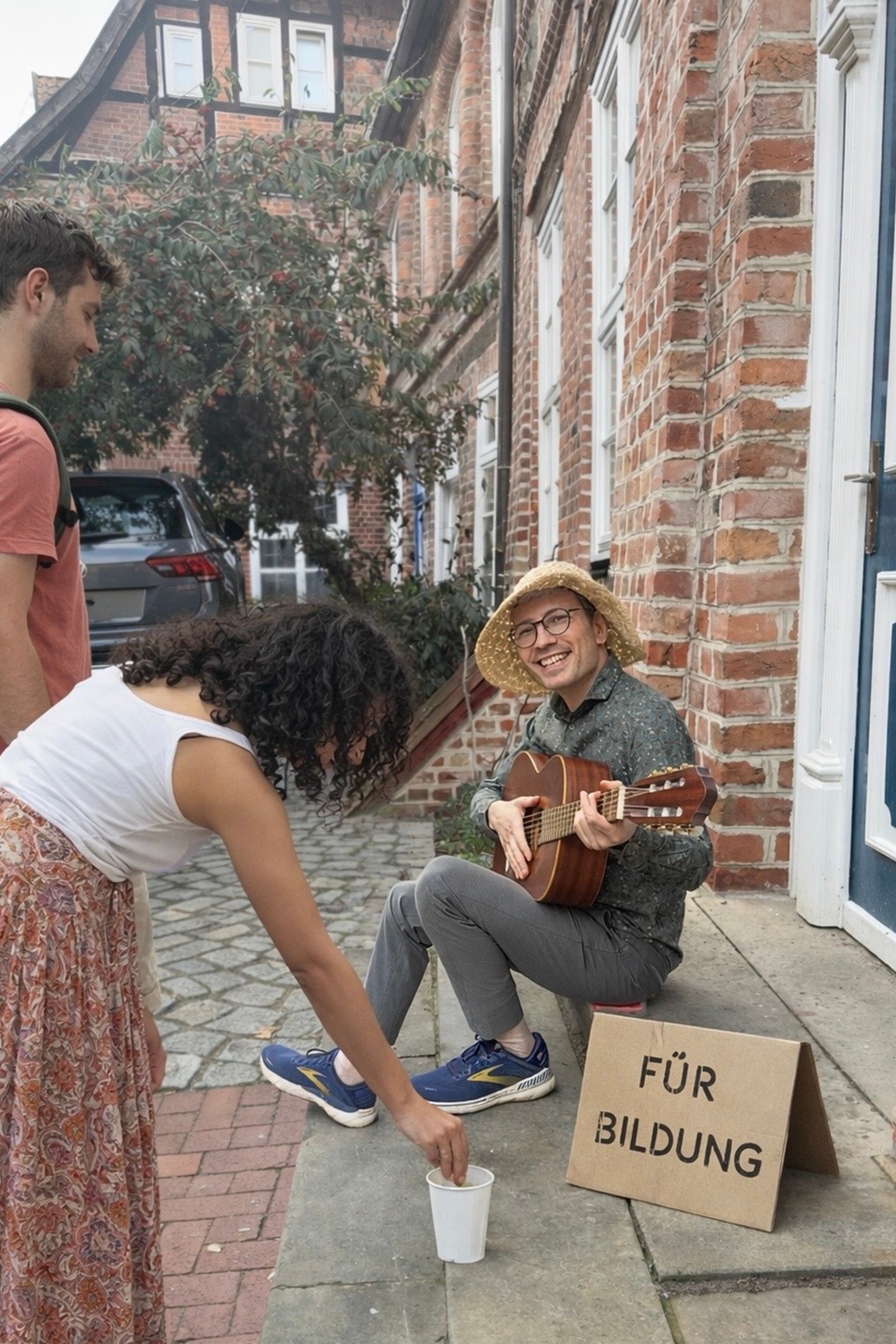 Der Lektorenausbilder Evgenij Unker als Straßenmusiker in der Lüneburger Altstadt: Er spielt Gitarre und singt und bekommt dafür Spenden von Passanten. KI-generiert.